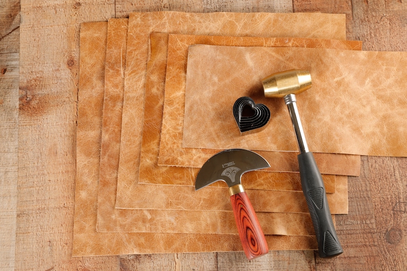 May include: A set of leather crafting tools including a leather cutting knife, a heart-shaped hole punch, and a brass hammer. The tools are laid out on a stack of brown leather sheets.