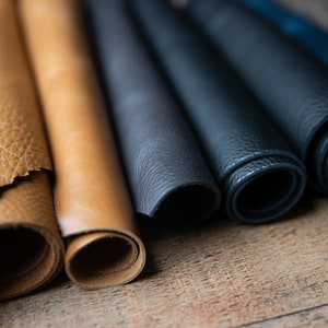 May include: Close-up of rolled up pieces of leather in various shades of brown and blue. The leather is arranged in a row on a wooden surface.