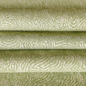 May include: A folded textile fabric featuring a light green base color with a white, swirling, topographical-like pattern. The material has a textured, woven appearance with visible horizontal folds creating depth across the surface of the cloth.