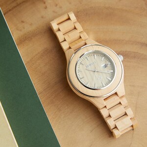 May include: A wooden wristwatch with a light brown band and a white face. The watch face has black hour and minute hands and a small black second hand.