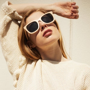 May include: A woman wearing a cream knitted jumper with a delicate open weave pattern. She is wearing large, dark sunglasses with a light wood frame.