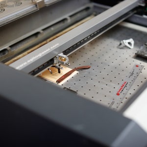 May include: A close-up of a laser cutter engraving a pair of glasses on a wooden surface. The laser cutter is a grey machine with a black and silver arm. The glasses are brown and are lying on a piece of wood. The machine has a ruler on the side and the text "www.apiloglaser.com" is visible on the machine.