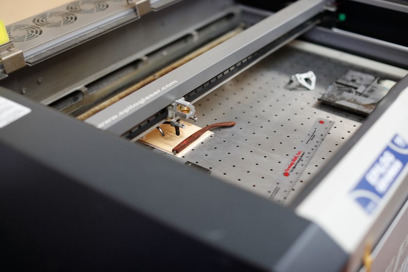 May include: A close-up of a laser cutter engraving a pair of glasses on a wooden surface. The laser cutter is a grey machine with a black and silver arm. The glasses are brown and are lying on a piece of wood. The machine has a ruler on the side and the text "www.apiloglaser.com" is visible on the machine.