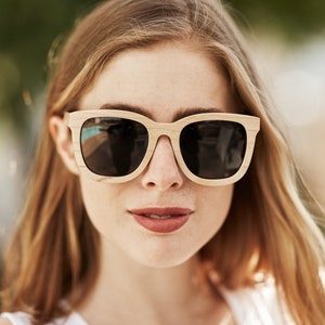 May include: A woman wearing sunglasses with a light brown wooden frame and dark lenses. The sunglasses have a square shape.