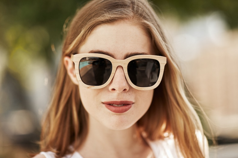 May include: A woman wearing sunglasses with a light brown wooden frame and dark lenses. The sunglasses have a square shape.
