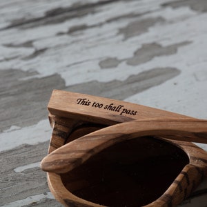 May include: Wooden sunglasses with a brown wood grain pattern. The sunglasses have a dark lens and the words "This too shall pass" are engraved on the arm.