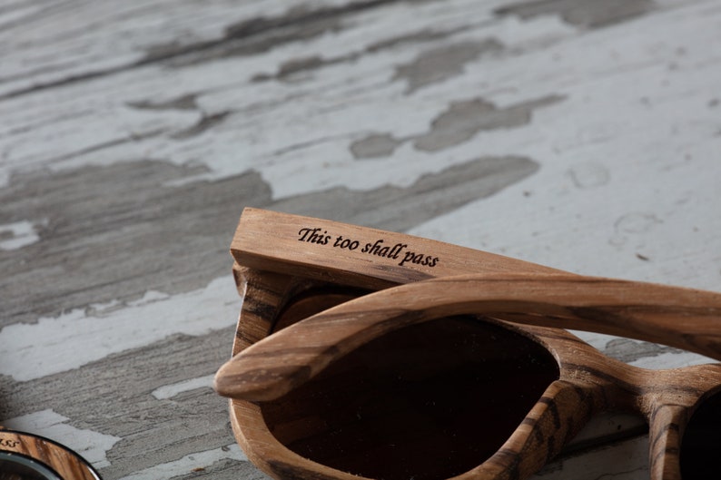 May include: Wooden sunglasses with a brown wood grain pattern. The sunglasses have a dark lens and the words "This too shall pass" are engraved on the arm.