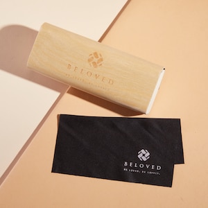 May include: A wooden glasses case with the text "BELOVED" and a logo of a stylised square with a cross inside. A black microfiber cleaning cloth with the same logo and text is next to the case.