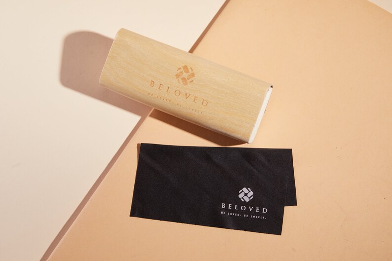 May include: A wooden glasses case with the text "BELOVED" and a logo of a stylised square with a cross inside. A black microfiber cleaning cloth with the same logo and text is next to the case.