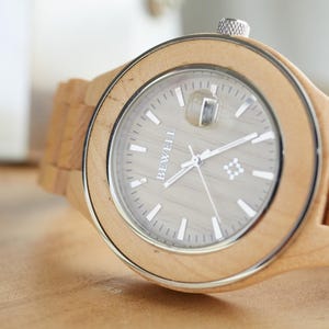 May include: A wooden wristwatch with a light brown band and a silver face. The watch face has a white background with black hands and the word "BEWELL" printed in white.