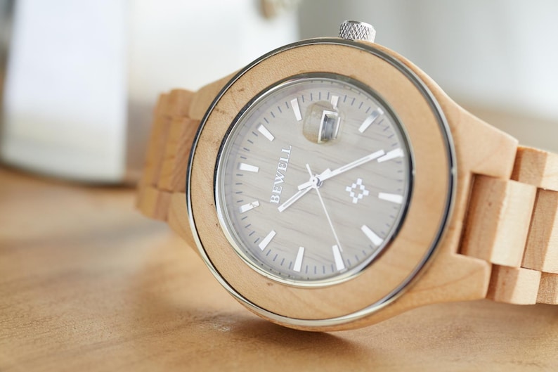 May include: A wooden wristwatch with a light brown band and a silver face. The watch face has a white background with black hands and the word "BEWELL" printed in white.