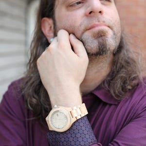 May include: A man wearing a maroon shirt with a patterned cuff, a wooden wristwatch with a light-colored face, and long brown hair.
