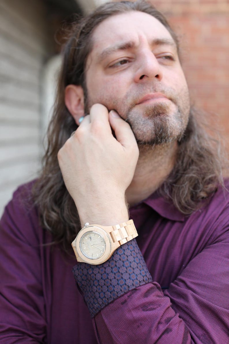 May include: A man wearing a maroon shirt with a patterned cuff, a wooden wristwatch with a light-colored face, and long brown hair.