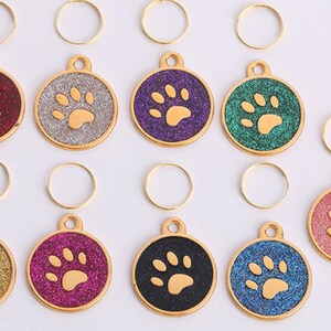 Op de afbeelding: Tien goudkleurige hondenpenning-bedels met een pootprintontwerp. Elke bedel heeft een andere gekleurde glitterachtergrond: rood, zilver, paars, groen, roze, goud, bordeauxrood, zwart, blauw en lichtroze. Elke bedel wordt geleverd met een gouden springring.