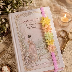 May include: A hardcover book titled "Jane Austen, Orgullo y Prejuicio" with a floral bookmark. The cover features a woman in a pink dress. A small candle, scissors, and flowers are also visible.