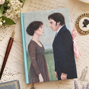 May include: A rectangular hardcover book featuring a painted illustration of a woman in a brown dress and a man in a dark blue coat facing each other. The book has a light blue spine, a pink ribbon bookmark, and a small silver heart-shaped charm.
