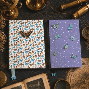 May include: Two rectangular notebooks with black spines. The left notebook features a white cover with a dense pattern of small blue and orange butterflies and a light blue ribbon bookmark. The right notebook has a purple cover with white stars and scattered blue butterfly accents, featuring a black ribbon bookmark with a small blue butterfly charm.