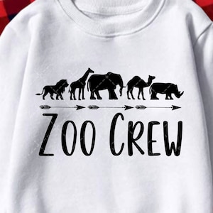 May include: White sweatshirt with black text that reads "Zoo Crew" and a line of black animal silhouettes including a lion, giraffe, elephant, rhino, camel, and zebra. The animals are facing right and there are arrows on either side of the animals.