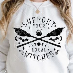 Puede incluir: Una sudadera blanca con un diseño gráfico negro que dice "Apoya a tus brujas locales". El gráfico presenta dos escobas cruzadas, un caldero, murciélagos y lunas.