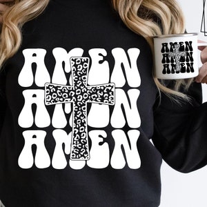 Op de afbeelding: Een zwart sweatshirt met een wit grafisch ontwerp van het woord "Amen" dat drie keer wordt herhaald met een luipaardprintkruis in het midden.