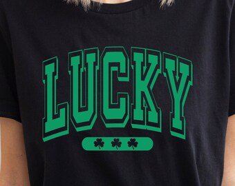 May include: Black t-shirt with green text that reads "LUCKY" in a varsity style font. Three green shamrocks are below the text.
