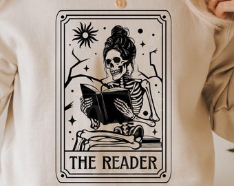 Può includere: Felpa beige con grafica in bianco e nero di uno scheletro che legge un libro, incorniciata da un bordo decorativo. Il testo "THE READER" è sotto l'immagine. Il logo Print Cuts SVG è nell'angolo in alto a sinistra.