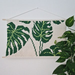 Puede incluir: Un tapiz de pared con un diseño tejido de hojas de monstera verdes sobre un fondo blanco. El tapiz está sujeto a una pared blanca con una cuerda blanca y listones de madera.