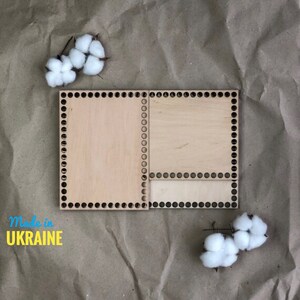 May include: Two wooden loom boards with holes around the edges, one is larger than the other. The boards are on a brown paper background with white cotton balls.