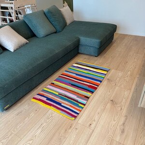 May include: A rectangular, handmade rug with a vibrant striped pattern. The rug features a yellow border and a colorful array of horizontal stripes in various colors, including red, blue, green, and pink. The rug is placed on a wooden floor.