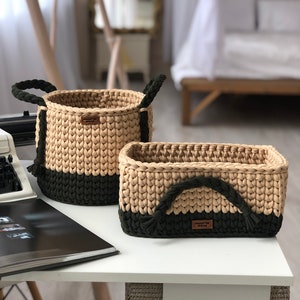 Puede incluir: Dos cestas de almacenamiento de crochet con rayas verdes y beige. Las cestas tienen asas gruesas de cuerda y una etiqueta de cuero marrón con la palabra "Handmade" en ella.