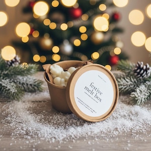 Könnte beinhalten: Eine festliche Schmelzbox mit Deckel, gefüllt mit weißen Wachsschmelzen, steht auf einer Holzoberfläche mit Kunstschnee. Der Deckel trägt den Text "festive melt box" und "Christmas Collection". Im Hintergrund ein Weihnachtsbaum mit Lichtern.