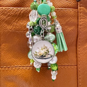 May include: A green and silver charm with a teapot and teacup design. The charm is attached to a key ring with a variety of green and white beads.