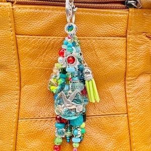 May include: A colorful beaded keychain with a silver clasp, featuring a variety of beads in shades of blue, green, red, and clear. A small silver hummingbird charm and a lime green tassel are also included. The keychain is hanging against a textured, tan leather background.