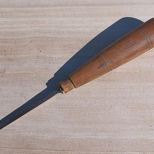May include: A hand tool featuring a dark, weathered wooden handle with a cylindrical shape and two incised rings near the top. Attached to the handle is a long, narrow, rectangular metal blade with a flat, squared-off tip and visible markings.