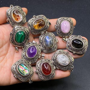 May include: A collection of ten rectangular silver-toned metal pendants, each featuring an ornate filigree border and a central oval cabochon gemstone. The stones include various colours such as green malachite, purple amethyst, iridescent abalone, and clear quartz.