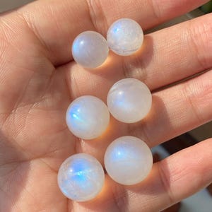 Op de afbeelding: Zes iriserende maansteen bollen, elk ongeveer 1,3 cm in diameter, worden in een hand gehouden. De bollen vertonen een melkachtige witte kleur met blauwe en paarse flitsen. De achtergrond is wazig.