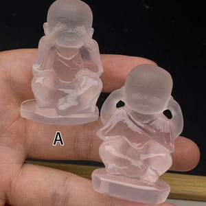 May include: Two translucent pink quartz Buddha statues. One statue has its hands covering its eyes, and the other has its hands covering its ears. Both are seated on a rectangular base. The statues are labelled A and B.