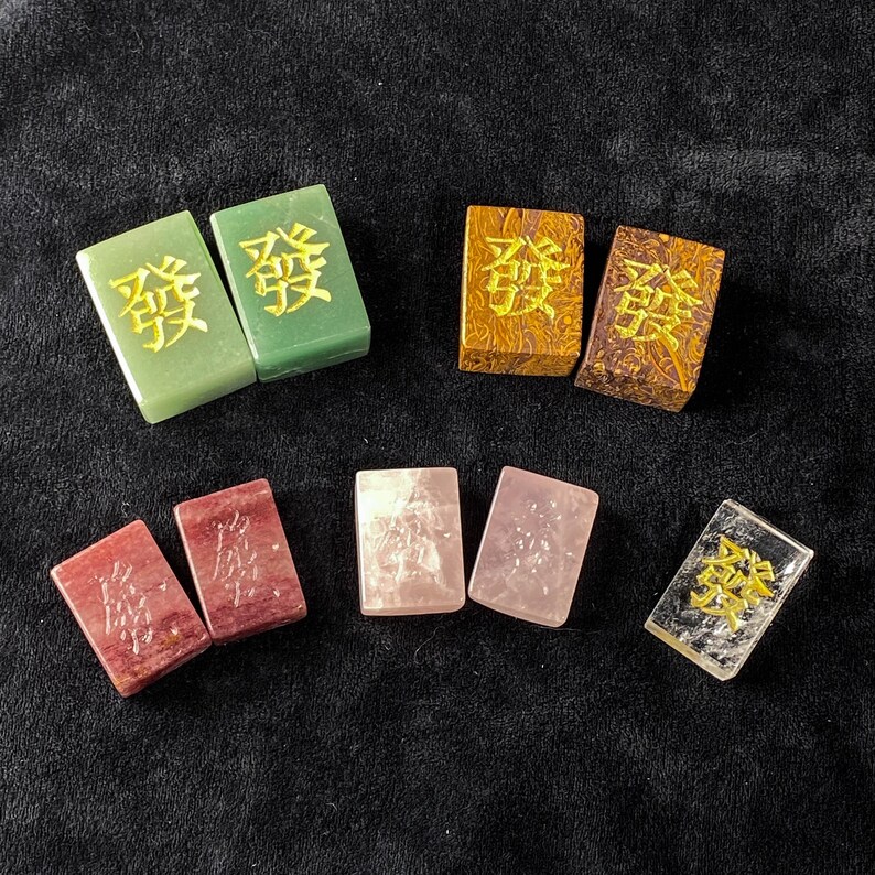 Pu&ograve; includere: Varie tessere di Mahjong colorate in tonalit&agrave; di verde, marrone, rosso, rosa e trasparente. Ogni tessera rettangolare presenta un carattere cinese in oro o giallo. Le tessere variano in consistenza e opacit&agrave;.