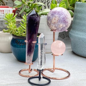 May include: An assortment of crystals and spheres on decorative metal stands. The crystals include a large amethyst point, a clear quartz crystal, and a rose quartz sphere. A larger sphere has a mottled purple and white pattern.