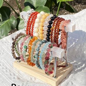 K&ouml;nnte beinhalten: Eine Pr&auml;sentation von Armb&auml;ndern mit Perlen in verschiedenen Farben, darunter Rot, Wei&szlig;, Schwarz und Rosa. Die Armb&auml;nder sind auf einem Display-St&auml;nder aus Holz und Metall angeordnet. Das Wort "Raw" ist unten im Bild in Wei&szlig; gedruckt.