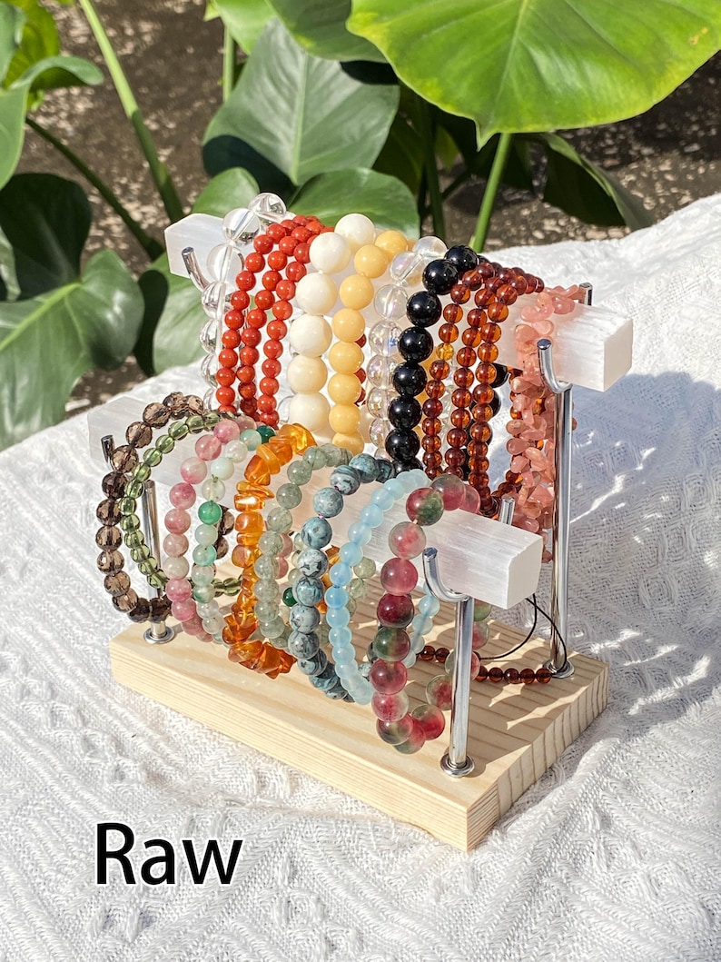 K&ouml;nnte beinhalten: Eine Pr&auml;sentation von Armb&auml;ndern mit Perlen in verschiedenen Farben, darunter Rot, Wei&szlig;, Schwarz und Rosa. Die Armb&auml;nder sind auf einem Display-St&auml;nder aus Holz und Metall angeordnet. Das Wort "Raw" ist unten im Bild in Wei&szlig; gedruckt.