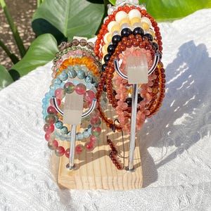 K&ouml;nnte beinhalten: Ein Holzst&auml;nder pr&auml;sentiert mehrere Armb&auml;nder mit Perlen in verschiedenen Farben, darunter Rot, Rosa, Gr&uuml;n und Blau. Die Armb&auml;nder bestehen aus verschiedenen Arten von Steinen und Perlen. Der St&auml;nder hat silberne Metallst&uuml;tzen und eine hellfarbige Holzbasis.