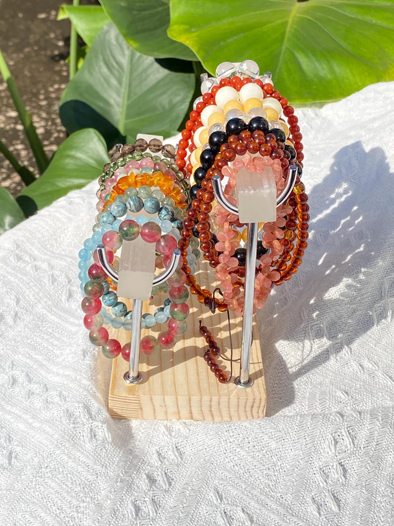 K&ouml;nnte beinhalten: Ein Holzst&auml;nder pr&auml;sentiert mehrere Armb&auml;nder mit Perlen in verschiedenen Farben, darunter Rot, Rosa, Gr&uuml;n und Blau. Die Armb&auml;nder bestehen aus verschiedenen Arten von Steinen und Perlen. Der St&auml;nder hat silberne Metallst&uuml;tzen und eine hellfarbige Holzbasis.