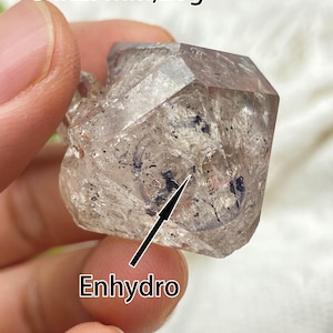 May include: A clear, faceted crystal with dark inclusions, held in a hand. The crystal measures 3.4x2.9cm and weighs 20g. The word "Enhydro" is written on the image.