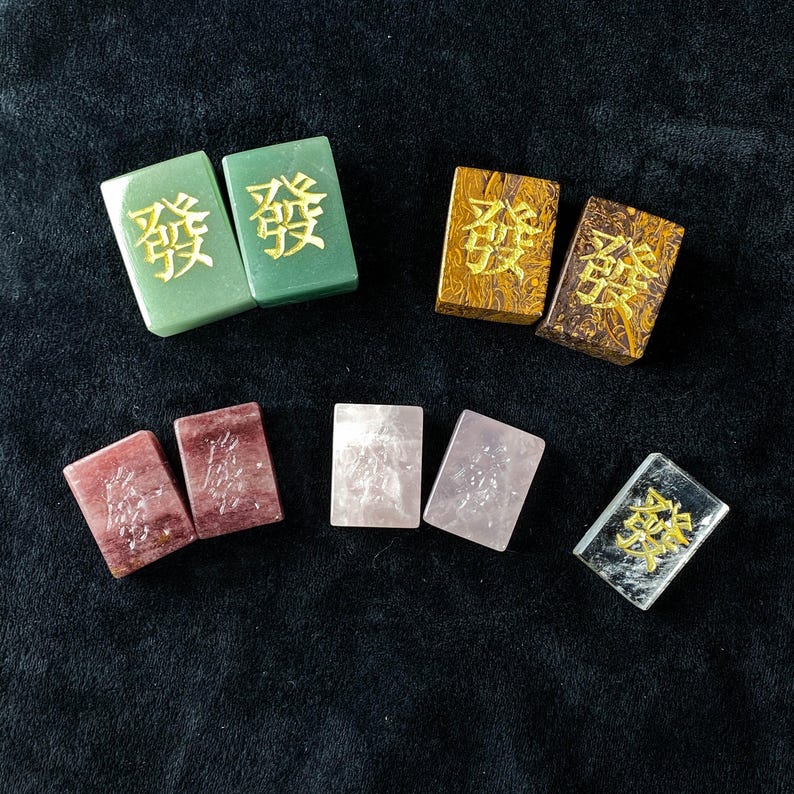 Pu&ograve; includere: Varie tessere di Mahjong in pietre preziose, verdi, marroni, rosa e trasparenti. Ogni tessera presenta caratteri cinesi dorati. Le tessere rettangolari sono disposte su uno sfondo scuro.
