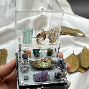 Op de afbeelding: Een heldere acryl display case met een verscheidenheid aan kleurrijke edelstenen en mineralen. De case is open en toont een selectie van kristallen, waaronder een blauwgroen kristal en een paarse, afgeronde steen. De case wordt in een hand gehouden.