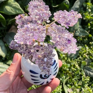 Puede incluir: Un árbol decorativo de piedras preciosas de amatista en una pequeña maceta de cerámica blanca con diseños de remolinos azules. El árbol tiene un tronco y ramas de alambre dorado, y las hojas están hechas de pequeñas piedras de amatista moradas. La maceta se sostiene en una mano.