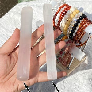 K&ouml;nnte beinhalten: Eine Hand h&auml;lt zwei Selenit-St&auml;be, einen polierten und einen rohen. Der polierte Stab ist zylindrisch mit einem abgerundeten Ende. Der rohe Stab ist rechteckig mit einer strukturierten Oberfl&auml;che. Der Text "Polished" und "Raw" sind mit Pfeilen beschriftet.