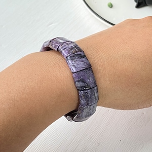 Puede incluir: Un brazalete de piedras preciosas de color morado y negro hecho con cuentas cuadradas. El brazalete está en la muñeca de una persona.