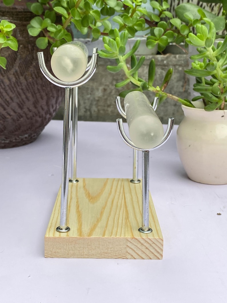 K&ouml;nnte beinhalten: Ein Holzst&auml;nder mit zwei transparenten, zylindrischen Objekten, die von silbernen Metallhaltern gehalten werden. Der St&auml;nder hat eine nat&uuml;rliche Holzmaserung. Gr&uuml;ne Pflanzen im Hintergrund.